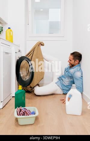 Smiling man sitting on the floor of a modern laundry room, playfully handling clothes in front of a washing machine, surrounded by detergent bottles a Stock Photo