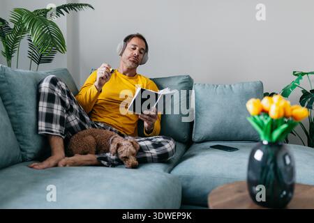 A man in pajamas sits comfortably on a sofa, wearing headphones and holding a notebook, with a cozy dog by his side. He enjoys a peaceful and relaxing Stock Photo