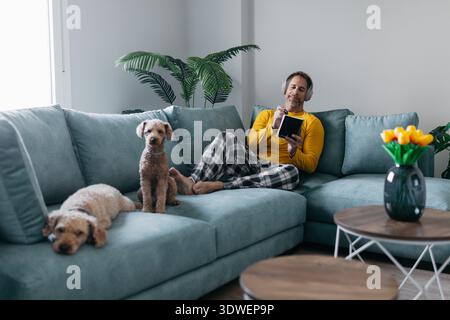 A man wearing a yellow shirt and plaid pajamas relaxes at home, sitting on a comfy couch with his dogs. He listens to music with headphones, enjoying Stock Photo