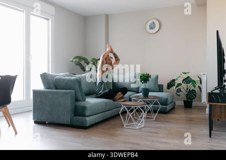 A man practices yoga at home, sitting cross legged on a comfortable sofa in the living room. The space is bright and inviting with plants and elegant Stock Photo