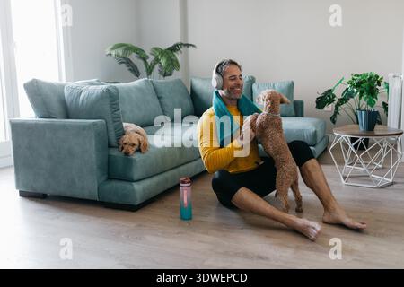 Man exercising at home, sitting on a wooden floor with his dog. Wearing headphones and a yellow shirt, he enjoys a workout session in a cozy, plant fi Stock Photo