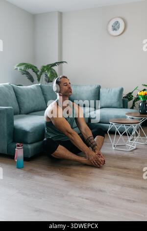 A man engages in a relaxing home workout session, stretching on the floor next to a comfortable teal sofa. He is wearing headphones and focusing on hi Stock Photo