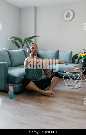 A man is practicing yoga exercises in his living room, sitting cross legged on the floor with eyes closed, and stretching his arms. The setting is cal Stock Photo