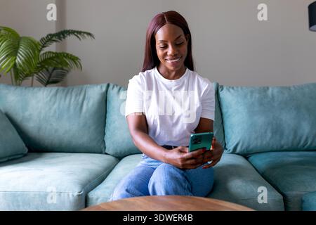 A woman sits comfortably on a teal sofa at home, smiling as she looks at her smartphone. She is wearing a white shirt and jeans, surrounded by a cozy Stock Photo