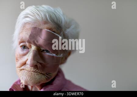 An elderly woman with white hair wears a facial wound. She is in a senior residence, and her expression is calm and thoughtful in a neutral setting. Stock Photo