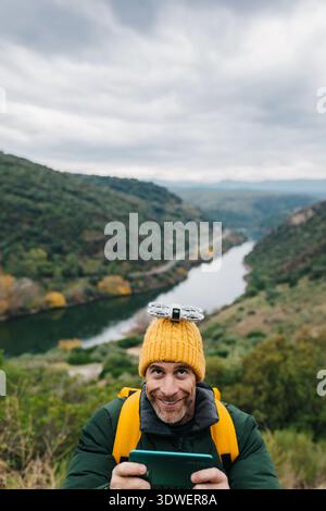 A man in a yellow hat smiles as he operates a drone in a beautiful, verdant mountainous landscape. The winding river and lush greenery create a breath Stock Photo