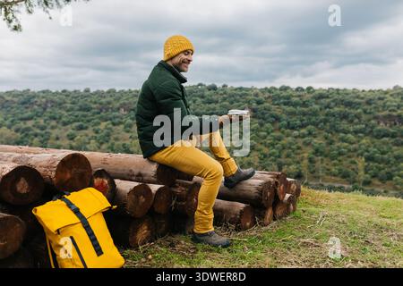 A man wearing a yellow beanie and green jacket is seated on stacked logs, smiling while holding a drone controller. The background features a lush, gr Stock Photo