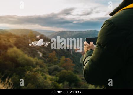 A man operates a drone using a handheld device amidst a picturesque landscape, highlighting the integration of technology and nature in capturing stun Stock Photo