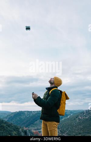 A man stands in a scenic mountainous landscape, focused on operating a small flying drone. The lush hills and expansive sky create a breathtaking back Stock Photo