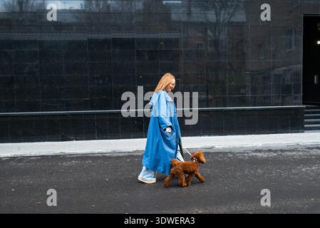 A woman in a blue coat walks her poodle along an urban street on a chilly autumn day. The dark, modern background contrasts with her vibrant outfit an Stock Photo