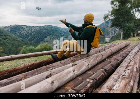 A man with a yellow backpack sits on wooden logs, controlling a drone in a picturesque landscape. He wears a matching yellow hat and enjoys the serene Stock Photo