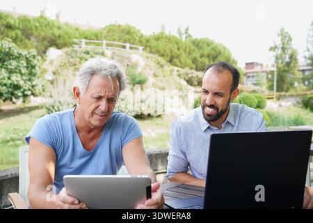 Two friends engage in a tech-savvy session at a table outside. One is focused on a tablet, while the other is working on a laptop. They enjoy a sunny Stock Photo