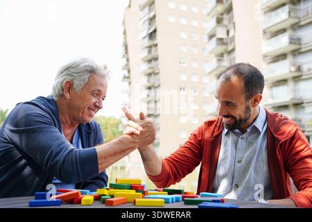 Two friends laughing and arm wrestling at a cafe table, surrounded by colorful blocks. They enjoy a sunny day, sharing fun and camaraderie in a casual Stock Photo