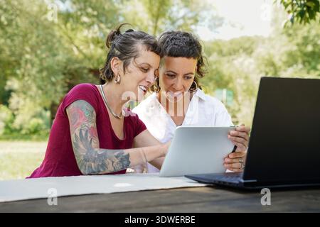 Two sisters enjoying a delightful day outdoors, engaging in digital activities. They are smiling and sharing information using a tablet, surrounded by Stock Photo