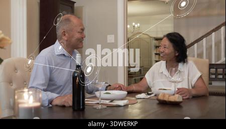 Holding hands Chinese couple at home table, wearing blue shirt, white blouse, wine bottle nearby Stock Photo