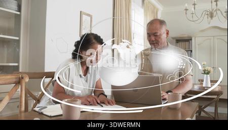 Reviewing papers, woman leaning while man checking laptop in home dining room, holographic globe Stock Photo