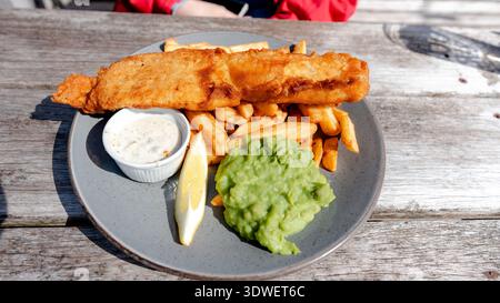 A classic dish of fish and chips is served with mushy peas, tartar sauce, and a wedge of lemon on a rustic wooden table, capturing the essence of Brit Stock Photo