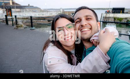 A smiling couple takes a joyful selfie by the sea in Cobh, Ireland. The background showcases the serene waterfront, creating a perfect setting for a m Stock Photo