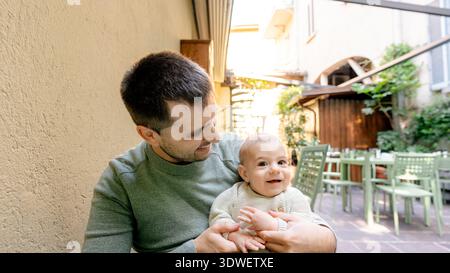 Smiling father holds his happy baby while sitting at a charming Italian cafe in Cernobbio. Sunlight filters through, creating a warm and joyful atmosp Stock Photo