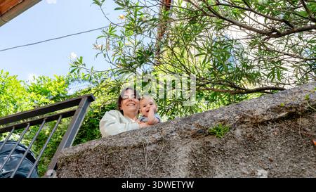A smiling woman holding a baby looks over a stone wall surrounded by lush greenery in Nesso, capturing a joyful moment on a sunny, clear-skied day. Stock Photo