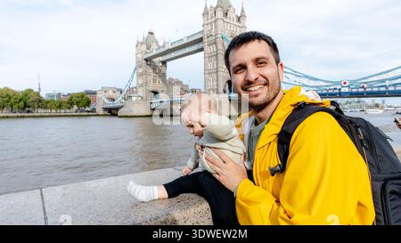 Happy family on a trip in London, England, with a parent holding a baby in front of the iconic Tower Bridge, showcasing a joyful moment on a beautiful Stock Photo