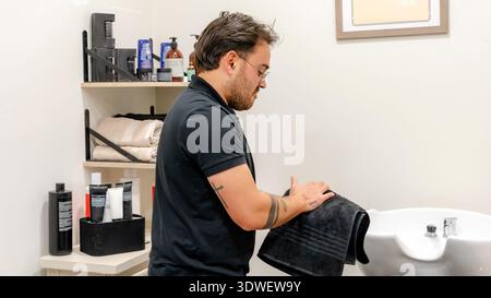 A professional barber meticulously dries his hands on a towel in a modern barbershop. Shelves in the background display various grooming products and Stock Photo
