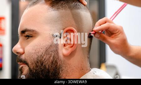 A close-up of a man getting a detailed haircut in a modern barbershop. A barber uses a precise tool to shape his hair, highlighting professional groom Stock Photo