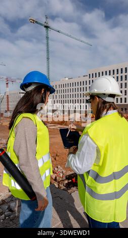 An architect and a technical engineer collaborate at a construction site, reviewing plans with a digital tablet. They wear safety gear, including helm Stock Photo