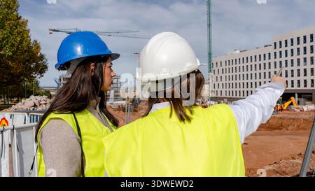 An architect and a technical engineer in hard hats and reflective vests discuss progress at a construction site. They stand amid cranes and building m Stock Photo