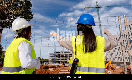 Female architect and technical engineer collaborate on a bustling construction site, overseeing progress while discussing plans. They wear safety gear Stock Photo