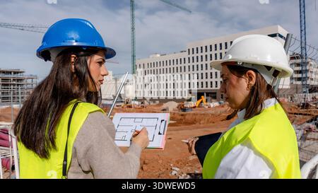 An architect collaborates with a technical engineer on a construction site, examining blueprints and discussing project details. Both wear safety gear Stock Photo