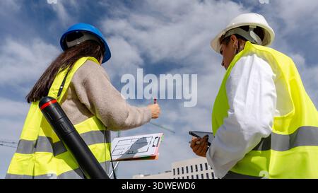 An architect and a technical engineer wearing safety gear discuss blueprints on a construction site. They analyze plans collaboratively, ensuring proj Stock Photo