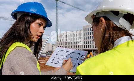 An architect and a technical engineer discuss building plans on a construction site. Wearing hard hats and safety vests, they review blueprints with c Stock Photo