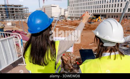An architect and a technical engineer in hard hats and safety vests collaborate on a construction site, reviewing plans and overseeing the progress of Stock Photo