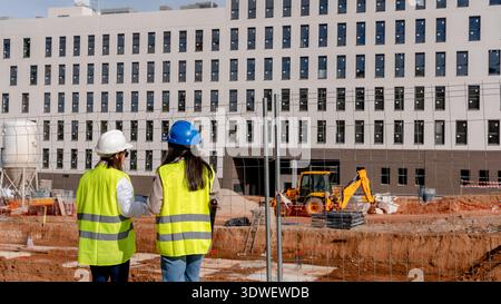 An architect and a technical engineer wearing safety vests and helmets stand on a construction site, discussing building plans with a partially constr Stock Photo