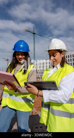 An architect and a technical engineer wearing safety vests and helmets are discussing project details on a construction site, using a tablet and docum Stock Photo