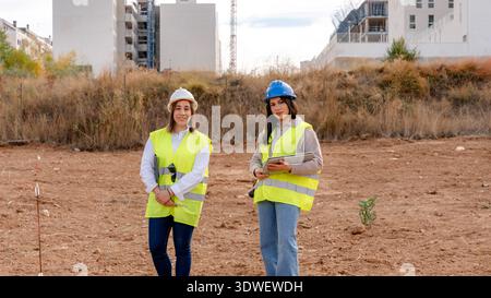Female architect and technical engineer inspecting a construction site. Dressed in high visibility vests and helmets, they hold plans and a laptop, fo Stock Photo