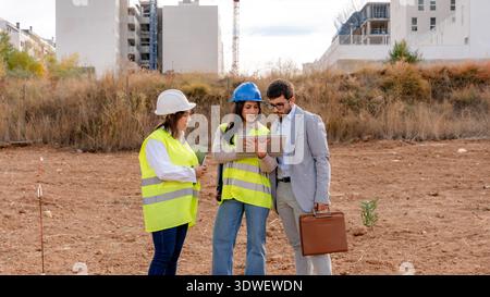An architect, a technical engineer, and an investor review plans on a construction site. They wear safety gear and collaborate on the project, demonst Stock Photo