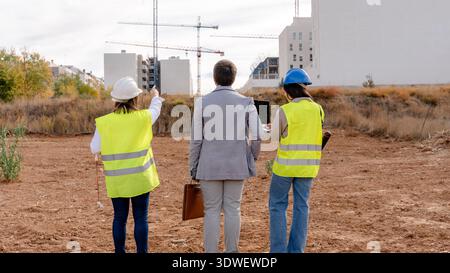 An architect, a technical engineer, and an investor stand together at a construction site, wearing safety gear and discussing plans for the future dev Stock Photo