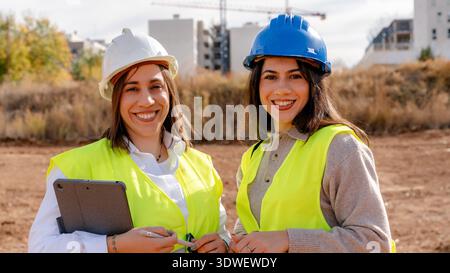 An architect and technical engineer at a construction site, wearing safety helmets and reflective vests, collaborate on project plans in an outdoor en Stock Photo