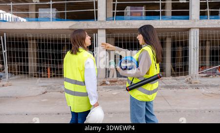 An architect and a technical engineer are engaged in discussion on a construction site, both wearing safety vests and holding helmets, exemplifying te Stock Photo