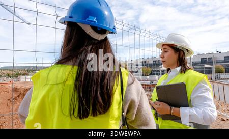 An architect and a technical engineer discuss plans on a construction site. Both are wearing safety helmets and high visibility vests, examining archi Stock Photo