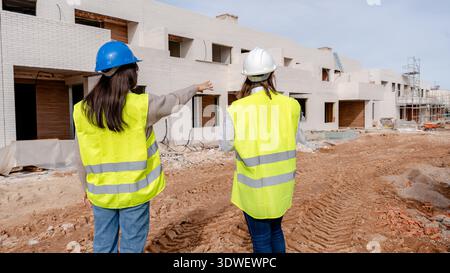 An architect and a technical engineer in high visibility vests and helmets inspect the progress of a residential construction site, analyzing the work Stock Photo