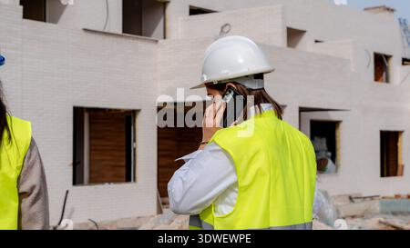 Architect and technical engineer on a construction site, wearing a hard hat and high visibility vest, engaging in a phone conversation while overseein Stock Photo