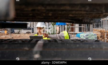 Female architect and technical engineer in protective gear collaborate on a construction site. They review plans and discuss project details amidst bu Stock Photo
