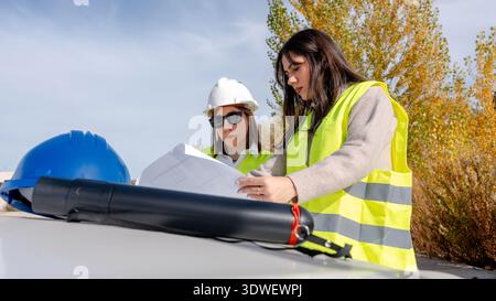 An architect and a technical engineer in safety vests and helmets collaborating and reviewing blueprints on a construction site surrounded by autumn f Stock Photo