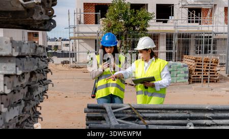 A female architect and technical engineer discuss plans on a construction site. They are wearing safety helmets and vests, emphasizing teamwork in a c Stock Photo