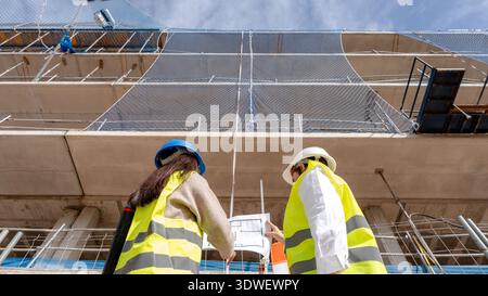 An architect and a technical engineer review blueprints on a construction site, collaborating on building plans. Both are wearing safety gear and obse Stock Photo