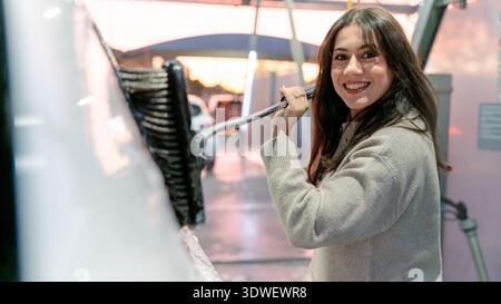 A woman with a smile cleans her car using a brush at a self-service car wash station, enjoying the efficient process of removing dirt with precision a Stock Photo