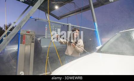 A woman smiles while using a high-pressure washer to clean her car at an outdoor self-service car wash. The setting sun casts a serene glow over the m Stock Photo